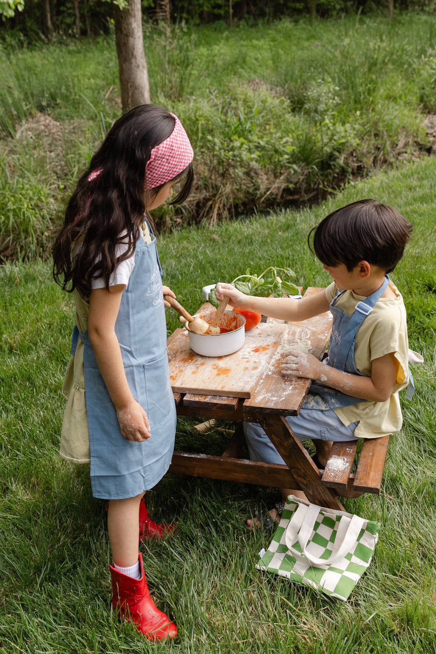 Kids wearing Little Chef Embroidered Linen cooking aprons outdoors cooking on wooden bench